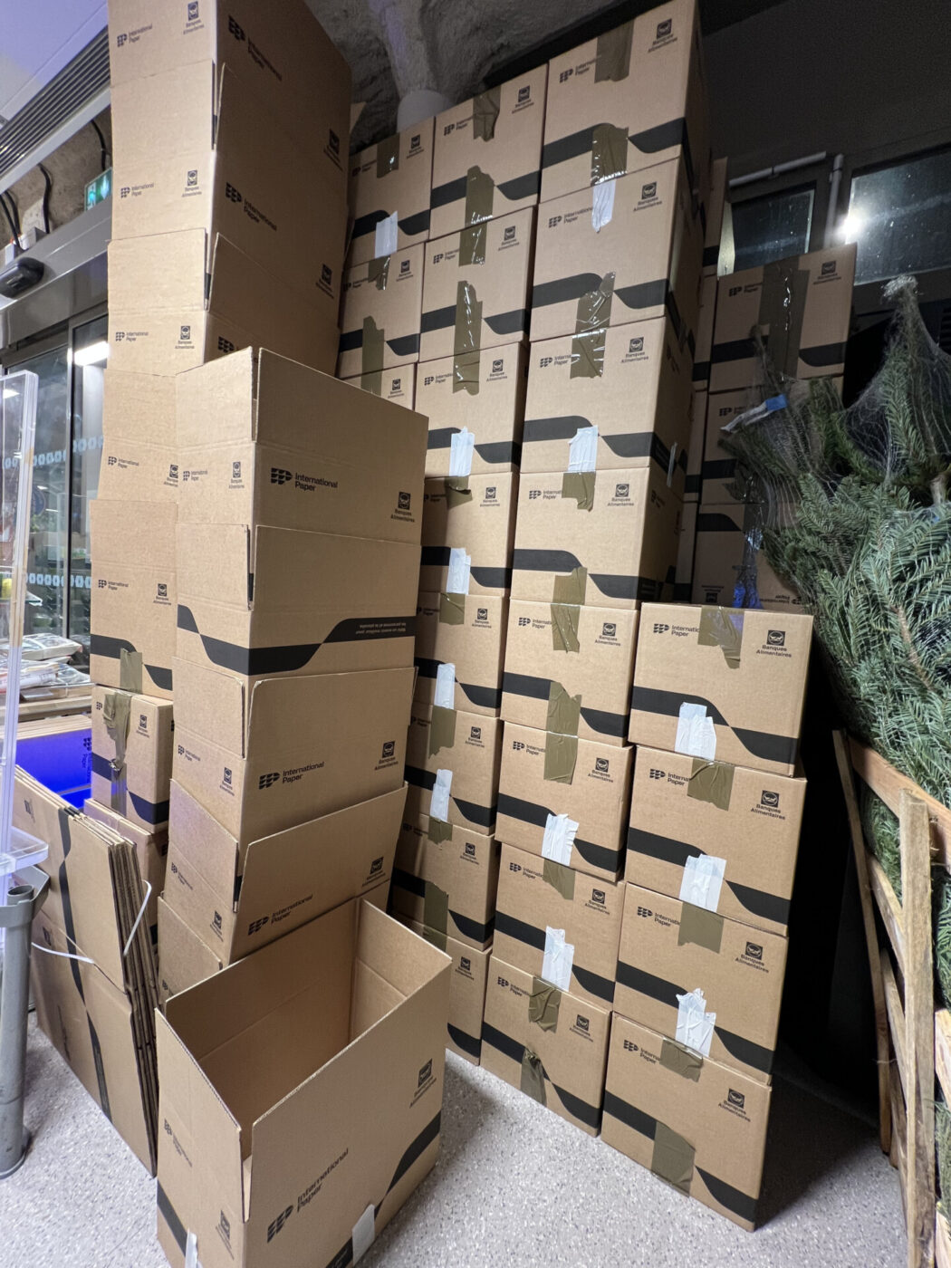 Une grande pile de boîtes en carton, certaines fermées et d'autres ouvertes, est disposée contre un mur à l'intérieur d'une pièce. À droite, on aperçoit partiellement des arbres à feuilles persistantes enveloppés. La scène suggère un stockage ou la préparation d'une collecte alimentaire par la Banque Alimentaire. - Rotary Club Paris Agora