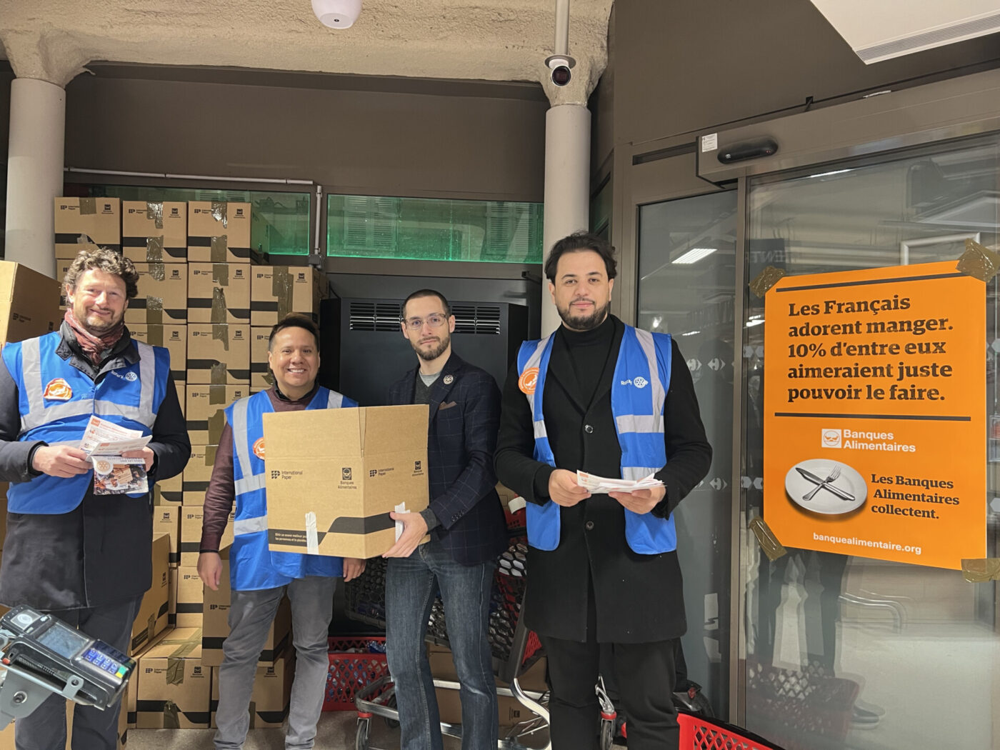 Quatre hommes se tiennent à l'intérieur, près d'une pile de cartons et d'un chariot, tenant des papiers et une boîte. Un panneau de collecte orange vif concernant l'aide alimentaire de la "Banque Alimentaire" est affiché sur la porte vitrée à côté d'eux. - Rotary Club Paris Agora