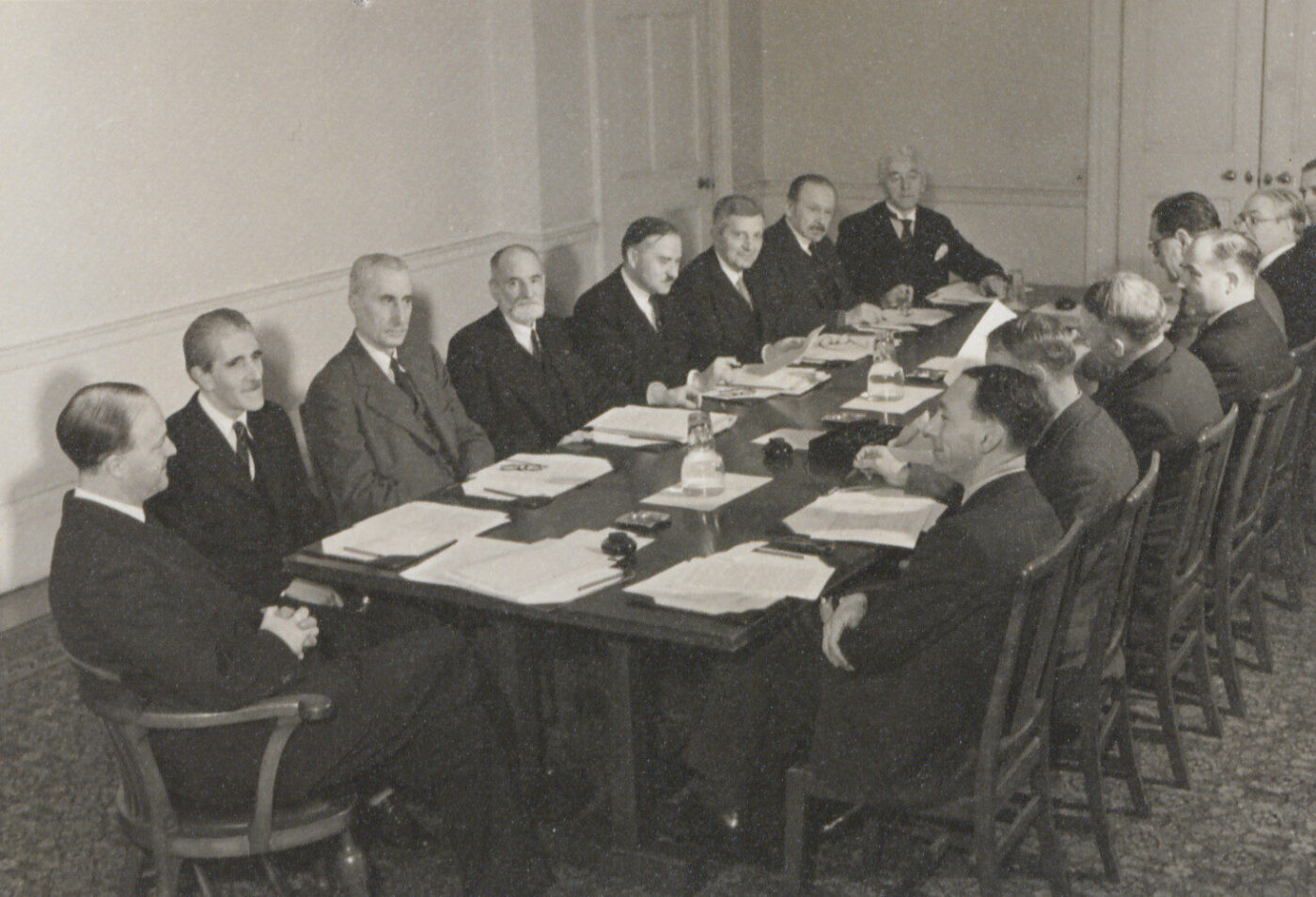 A group of men in suits sit around a large rectangular table in a formal meeting room, discussing documents. The setting evokes Rotary's role in the creation of UNESCO, with plain walls and a closed door in the background. - Rotary Club Paris Agora
