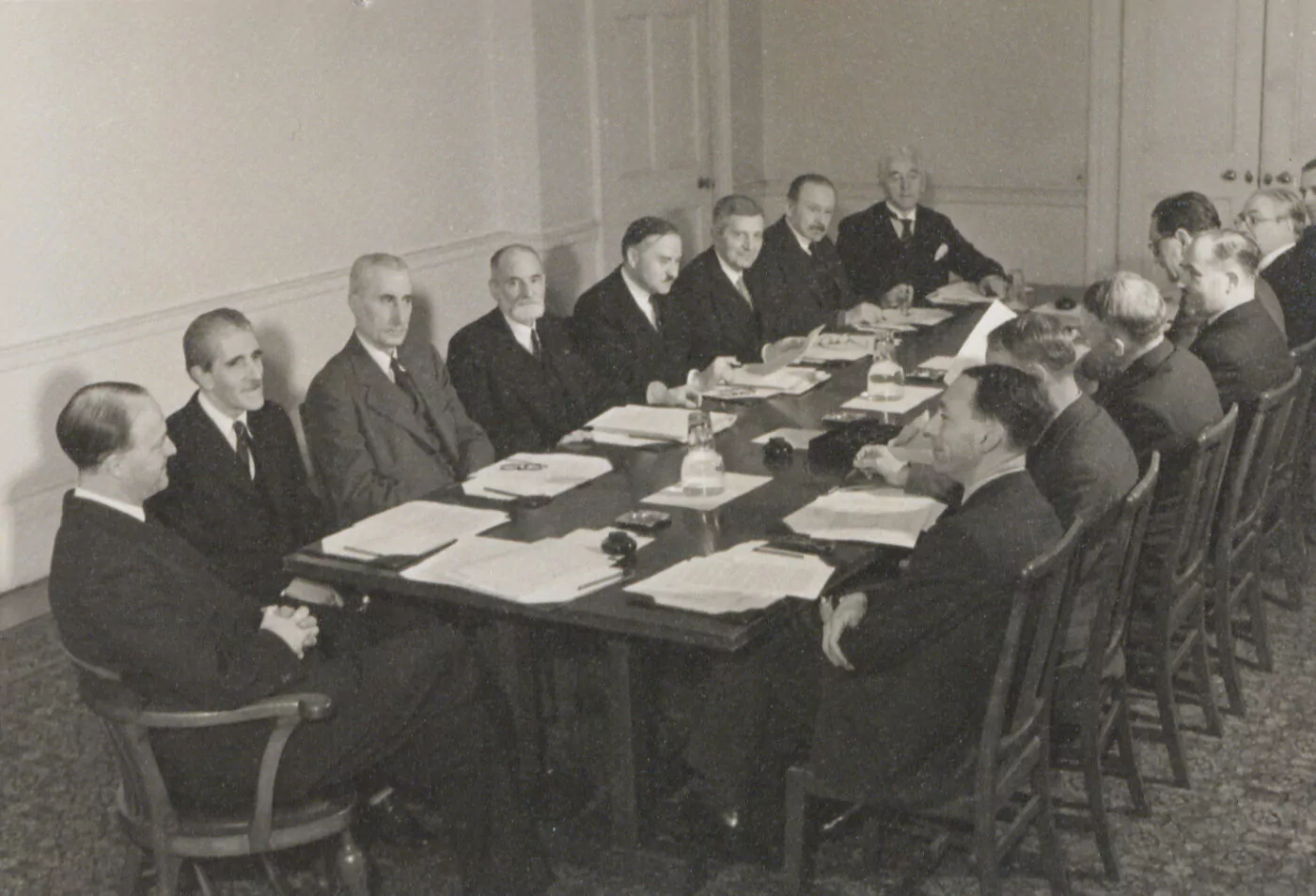 A group of men in suits sit around a large rectangular table in a formal meeting room, discussing documents. The setting evokes Rotary's role in the creation of UNESCO, with plain walls and a closed door in the background. - Rotary Club Paris Agora