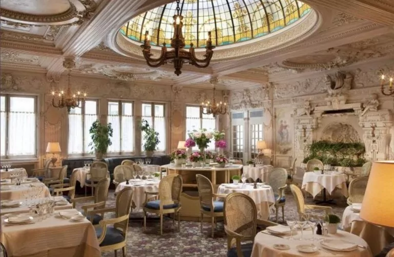 Élégante salle à manger de restaurant avec des plafonds ornés, des lustres, une grande lucarne en verre teinté et des sculptures murales classiques. Les tables sont dressées avec du linge blanc et un centre de table floral décore le centre de la pièce. - Rotary Club Paris Agora