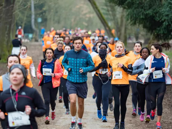 Un groupe de coureurs, dont certains portent des chemises orange, participent à une course sur le sentier d'un parc. Les participants semblent concentrés alors qu'ils courent au milieu des arbres par un temps couvert. Les dossards sont visibles sur leurs chemises. - Rotary Club Paris Agora