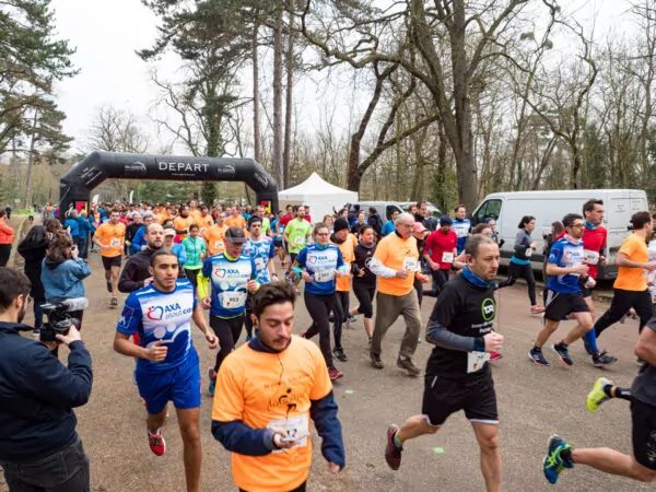 Un groupe de coureurs participe à une course, portant des dossards numérotés et des chemises colorées, alors qu'ils passent sous une arche gonflable noire marquée "DEPART" ; des arbres et des véhicules de soutien sont visibles à l'arrière-plan. - Rotary Club Paris Agora