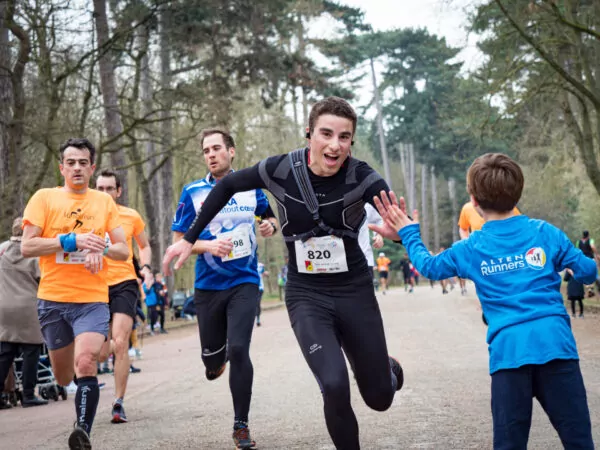 Un coureur portant une tenue noire et le dossard 820 sourit et donne la main à un enfant vêtu d'un maillot bleu, tandis que d'autres coureurs vêtus de maillots orange et bleus le suivent sur un sentier bordé d'arbres. - Rotary Club Paris Agora
