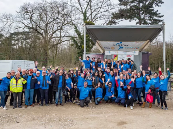 Un grand groupe de personnes, dont beaucoup portent des vestes bleues, posent et saluent pour une photo devant une scène avec une bannière "La Solirun" dans un cadre extérieur avec des arbres et des véhicules à l'arrière-plan. - Rotary Club Paris Agora