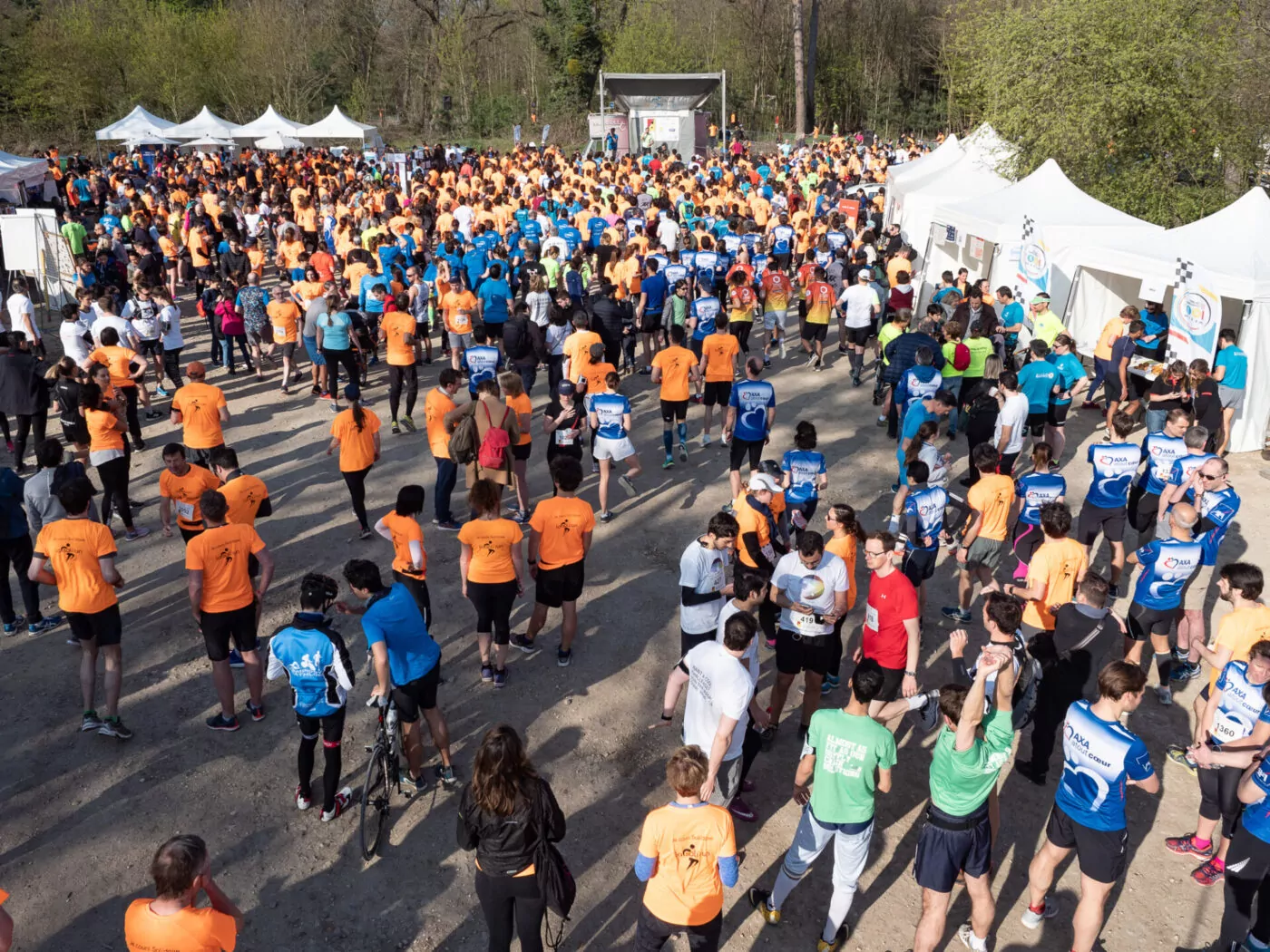 Une grande foule de personnes vêtues de chemises de sport colorées se rassemblent en plein air près de tentes blanches, se préparant à une course ou à un événement ; certains s'étirent, marchent ou discutent sous un ciel ensoleillé. - Rotary Club Paris Agora
