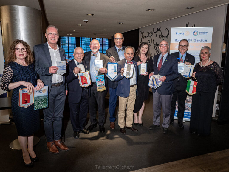 Onze personnes se tiennent à l'intérieur, en tenue de soirée, tenant diverses bannières et objets du Rotary. Soulignant les relations internationales, un présentoir du Rotary International est visible à l'arrière-plan, ainsi qu'une fenêtre et quelques meubles. - Rotary Club Paris Agora