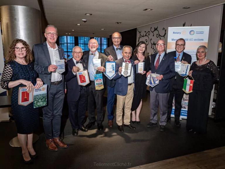 Eleven people stand inside, dressed in formal attire, holding various Rotary banners and items. Highlighting international relationships, a Rotary International display is visible in the background, along with a window and some furniture. - Rotary Club Paris Agora