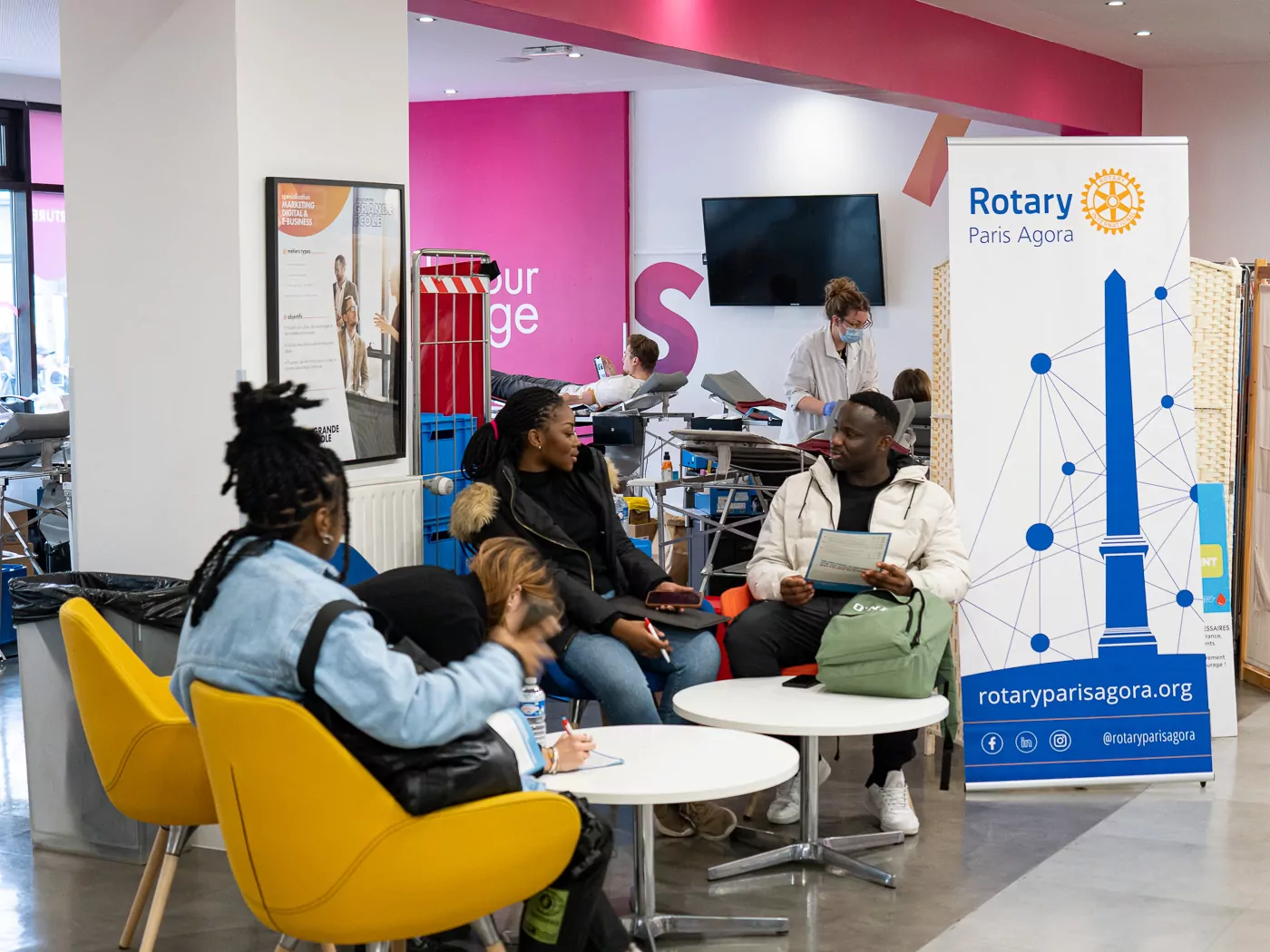 Quatre personnes sont assises et discutent dans une salle d'attente aux chaises jaune vif. Derrière elles, une banderole du Rotary Paris Agora met en avant la collecte de sang. Le personnel médical s'occupe des patients qui font un don dans le cadre de Mon sang pour les autres, dans une salle moderne et bien éclairée. - Rotary Club Paris Agora