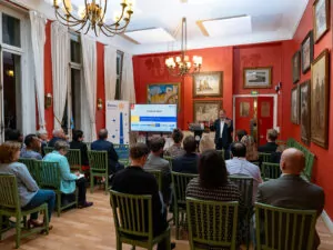 A group of people are sitting on green chairs facing a speaker at the front of a room with red walls, paintings, chandeliers, and a screen displaying a Rotary Club presentation on polio eradication. - Rotary Club Paris Agora
