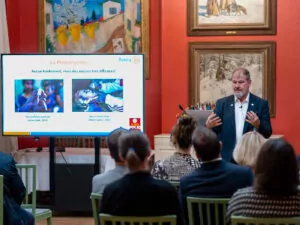 A man stands and addresses an audience in a room with painted walls. A slide presentation on polio vaccines, featuring photos of children and text in French, is displayed on a large screen next to him, highlighting the fight against polio. - Rotary Club Paris Agora