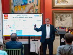 A man in a suit stands and gestures in front of a screen displaying a map of Pakistan and Afghanistan, discussing recent polio outbreaks with an audience sitting attentively in the conference room. - Rotary Club Paris Agora