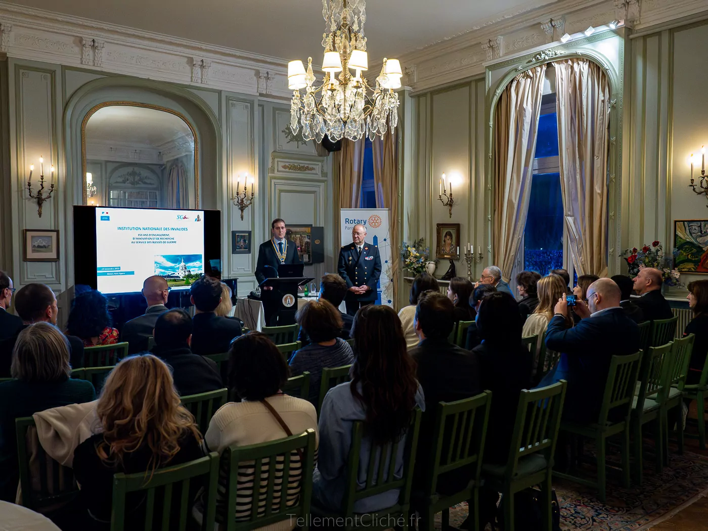 A group of people attends a presentation in a solemn hall of the National Invalides Institute with chandeliers, green chairs, and ornate walls. Two men stand at the front, near a projection screen displaying a Rotary banner. - Rotary Club Paris Agora