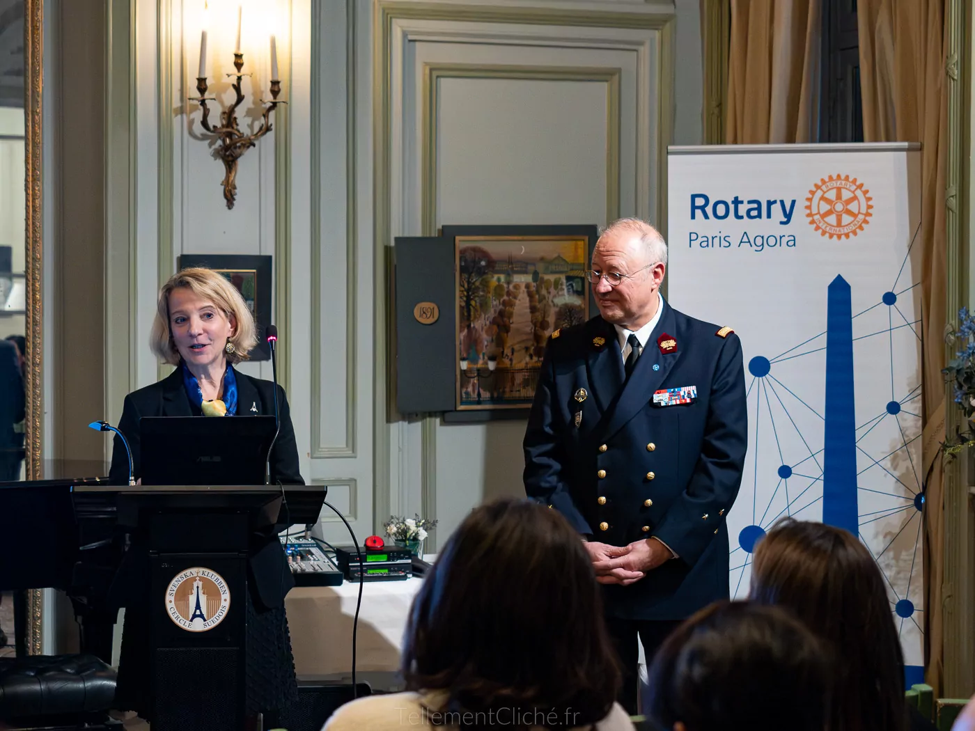 Une femme s'exprime sur un podium tandis qu'un homme en uniforme se tient à ses côtés à l'Institution nationale des Invalides à Paris. Derrière eux se trouve une bannière du Rotary Paris Agora et un public est assis face à eux dans une salle officielle. - Rotary Club Paris Agora