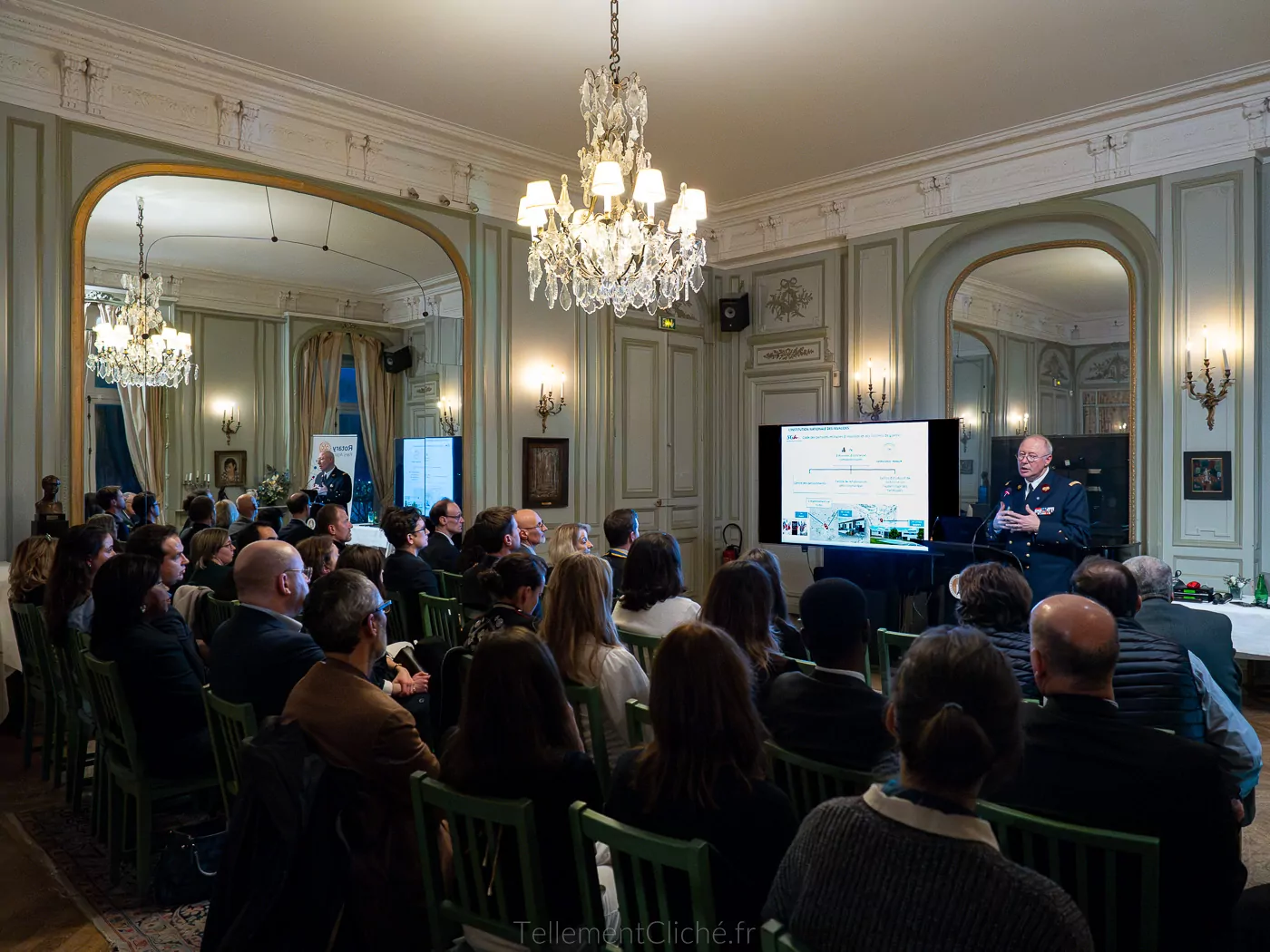 Un homme en uniforme fait une présentation à un public assis dans les salles ornées de l'Institution nationale des Invalides, avec des lustres, des miroirs et un grand écran affichant des informations. Les participants font face au présentateur et l'écoutent attentivement. - Rotary Club Paris Agora