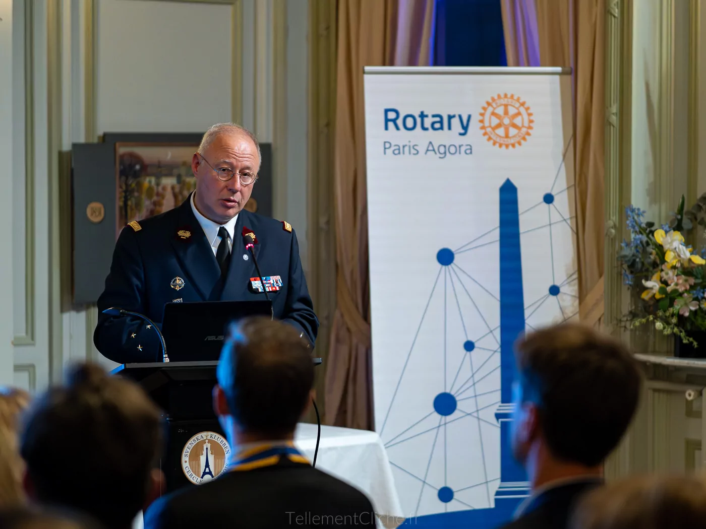 A man in military uniform speaks at a podium in front of an audience in Paris. Behind him, a banner with the logo of Rotary Paris Agora, the Eiffel Tower, network lines, and references to the National Invalides Institution. - Rotary Club Paris Agora