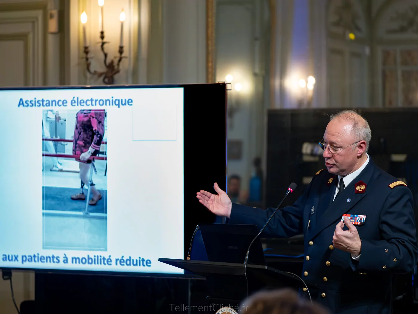 Lors d'une conférence à Paris, un homme en uniforme militaire de l'Institution nationale des Invalides présente l'"Assistance électronique aux patients à mobilité réduite", en montrant une diapositive montrant une personne utilisant un déambulateur. - Rotary Club Paris Agora