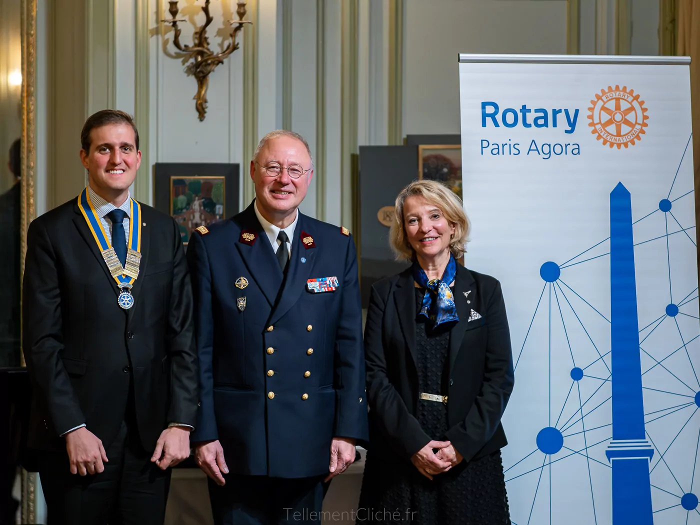 Trois personnes posent à l'intérieur près d'une banderole du Rotary Paris Agora. L'homme de gauche porte une médaille, celui du centre est en uniforme militaire de l'Institution nationale des Invalides et la femme de droite est vêtue de noir. - Rotary Club Paris Agora