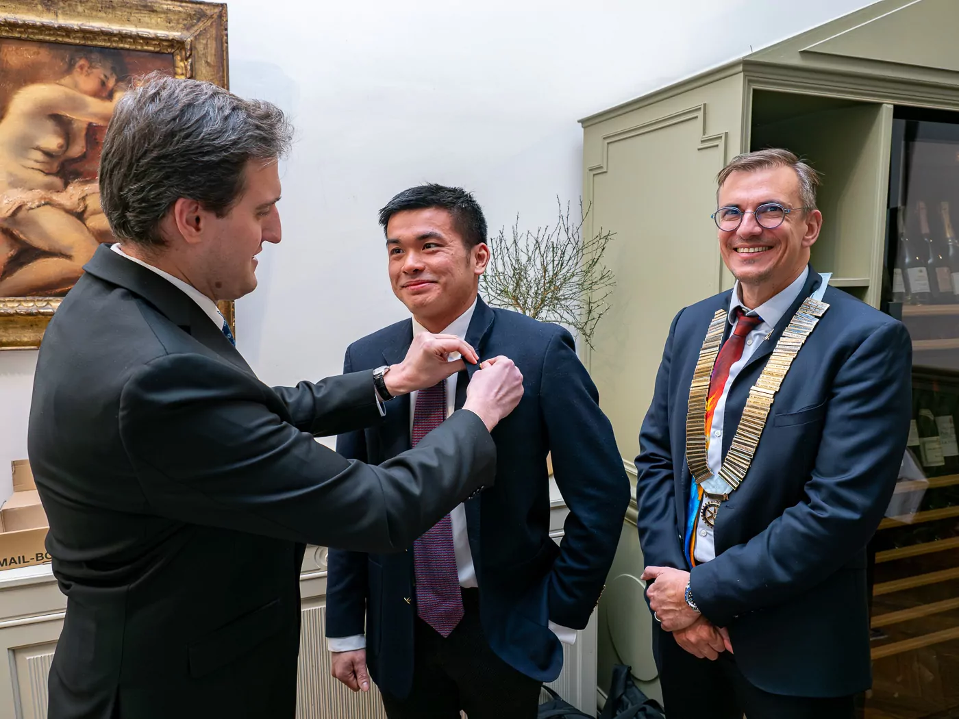 Governor Alexandre Guiard fastens a badge onto another man's jacket, while a third, wearing a ceremonial chain, smiles approvingly in congratulations. A painting hangs on the wall behind them. - Rotary Club Paris Agora