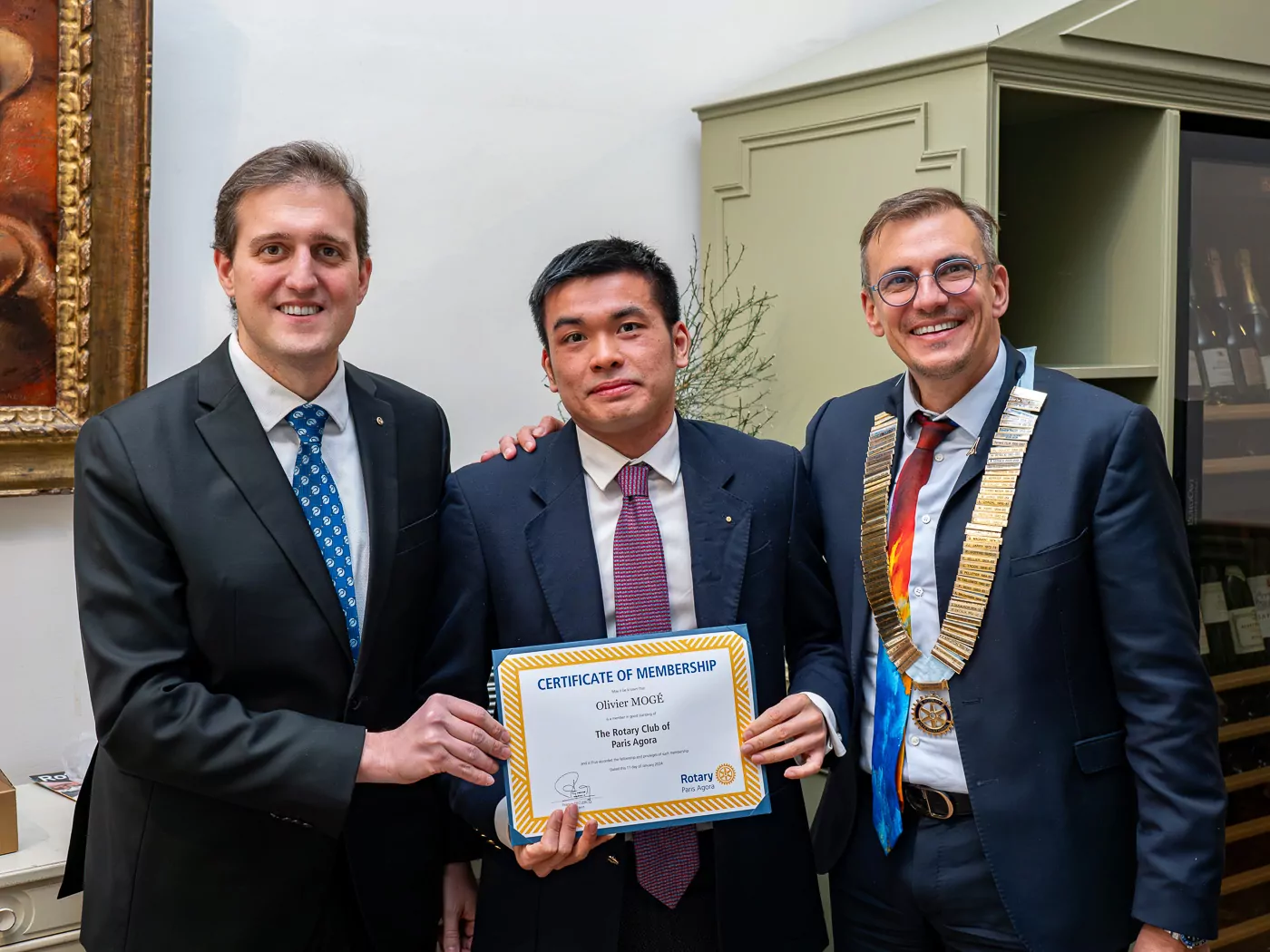 Three men in suits stand inside. Governor Alexandre Guiard is in the center and holds a framed membership certificate. The two men on either side of him smile in congratulations, and the man on the right wears a large medal and chain. - Rotary Club Paris Agora