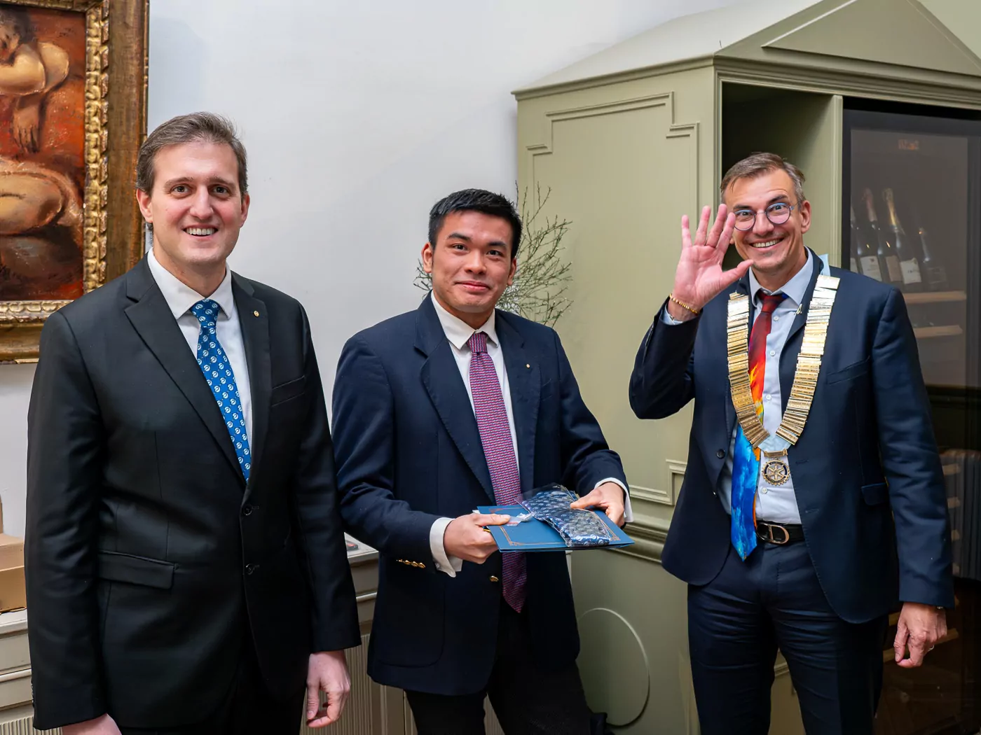 Three men in suits are standing inside. Governor Alexandre Guiard, on the right, is wearing a large ceremonial gold chain and making an "OK" gesture. The man in the middle is holding a book. All three men are looking at the camera and smiling, their expressions clearly conveying their congratulations. - Rotary Club Paris Agora