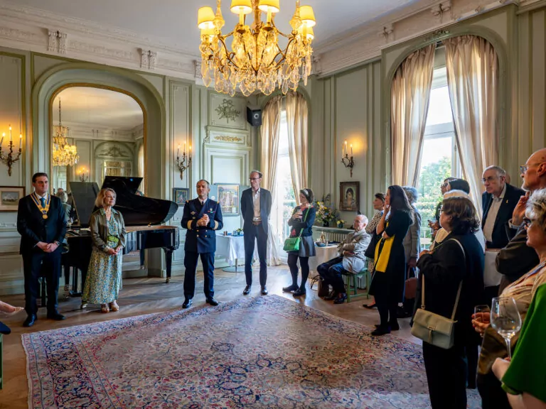 A group of people stands listening attentively in an elegant hall at Les Invalides, adorned with chandeliers, large windows, and a grand piano. It's a refined musical evening where some guests hold drinks while others are seated. The setting appears formal and well-lit. - Rotary Club Paris Agora