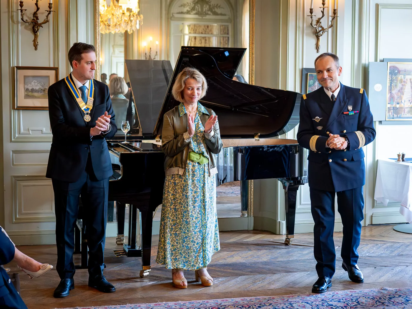 At a musical evening at Les Invalides, three people stand before a grand piano: the man on the left is wearing a suit with a medallion, the woman in the center is wearing a floral dress and jacket, and the man on the right is in a military uniform. They are all applauding. - Rotary Club Paris Agora