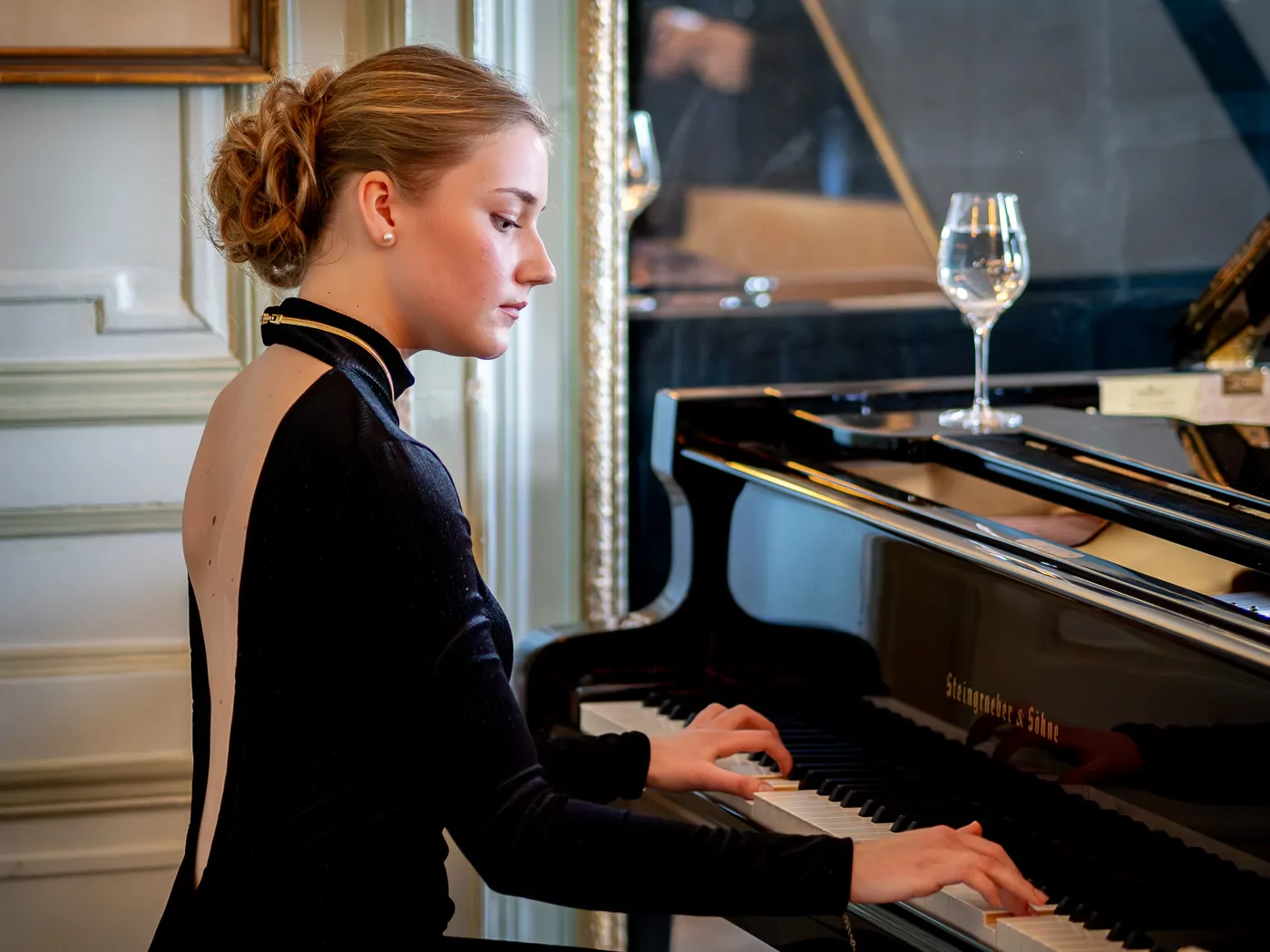 A woman in a black dress plays a grand piano in an elegant room, creating the ambiance of a musical evening. A wine glass rests on the piano, and a mirror reflects part of the scene as the woman concentrates on the keys. - Rotary Club Paris Agora
