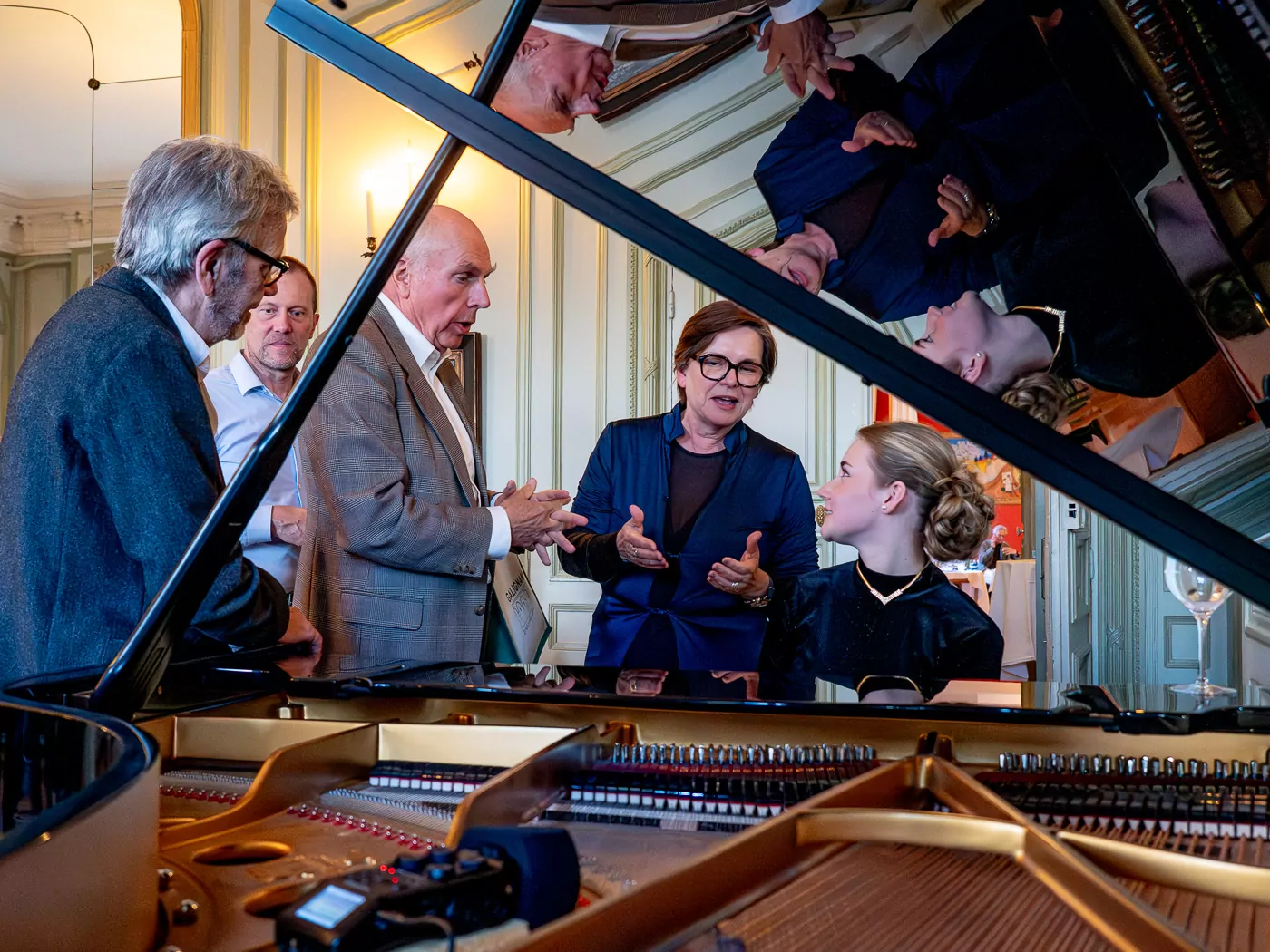 Five adults stand and chat around a grand piano in an elegant room during a musical evening. Their reflections shimmer on the open lid, which dominates the foreground, revealing the piano's strings, a microphone, and echoes of the sophistication of the Invalides. - Rotary Club Paris Agora