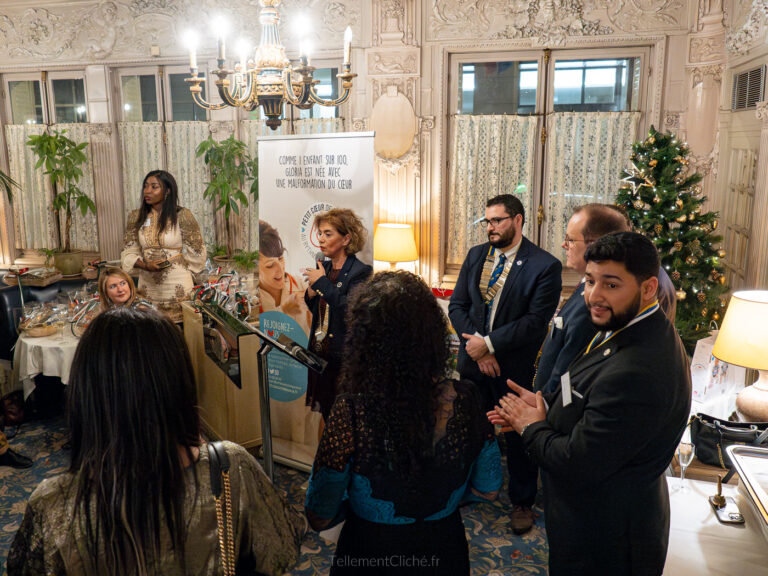 Un groupe de personnes habillées de manière formelle se réunit dans une salle décorée d'un lustre, d'un arbre de Noël et d'une table de buffet. Lors de la Soirée de Noël Interclub, une femme prend la parole sur un podium tandis que d'autres personnes écoutent debout lors de cet événement festif. - Rotary Club Paris Agora