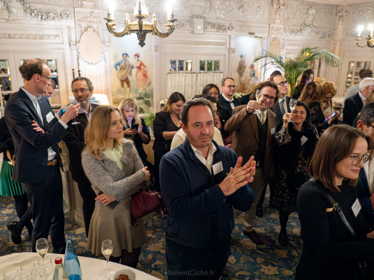 Un groupe de personnes, vêtues de tenues d'affaires ou de soirée, se tient debout et discute lors d'un événement Interclub dans une salle ornée de murs et de lustres décoratifs. Certains applaudissent, d'autres tiennent un verre ou utilisent leur téléphone. - Rotary Club Paris Agora