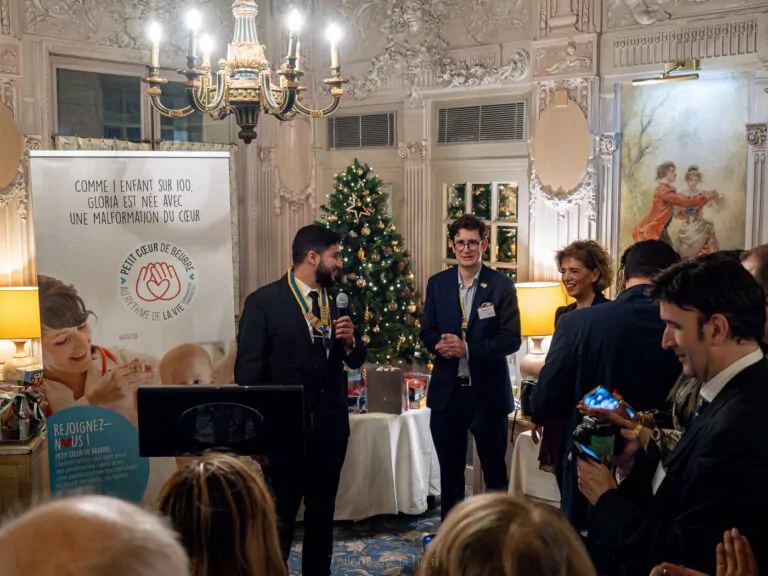 Un homme s'exprime sur un podium dans une salle décorée d'un sapin de Noël et de lustres lors d'une Soirée de Noël de l'Interclub. Des invités en tenue de soirée écoutent attentivement, tandis qu'une bannière sur les malformations cardiaques souligne l'objectif de l'événement. - Rotary Club Paris Agora