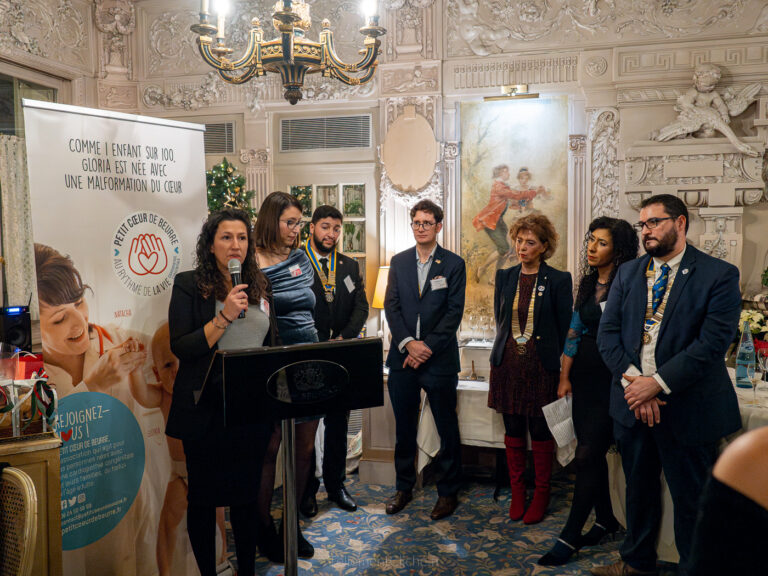 Une femme parle sur un podium tandis que six personnes se tiennent à ses côtés dans une salle décorée lors d'une Soirée de Noël de l'Interclub. Une bannière sur les malformations cardiaques, un lustre, des murs ornés et un tableau sont visibles à l'arrière-plan. - Rotary Club Paris Agora