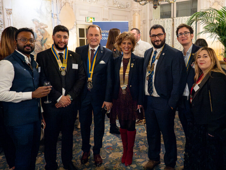 Un groupe de sept adultes, cinq hommes et deux femmes, habillés de manière formelle ou semi-formelle, se tiennent debout et posent à l'intérieur lors d'une soirée Interclub. Plusieurs d'entre eux portent des médailles et des insignes, ce qui laisse supposer qu'il s'agit d'un événement cérémoniel. Une personne tient un verre à vin. - Rotary Club Paris Agora
