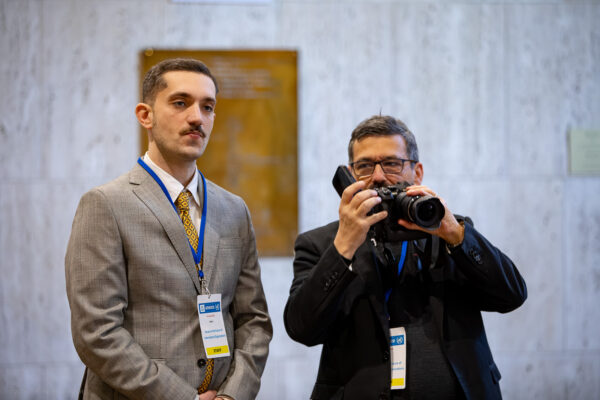 Deux hommes portant des badges de conférence se tiennent à l'intérieur ; l'un en costume gris avec une expression sérieuse, l'autre en costume noir tenant un appareil photo et capturant ce rendez-vous historique pour le multilatéralisme. Un mur de marbre et une plaque sont visibles à l'arrière-plan. - Rotary Club Paris Agora