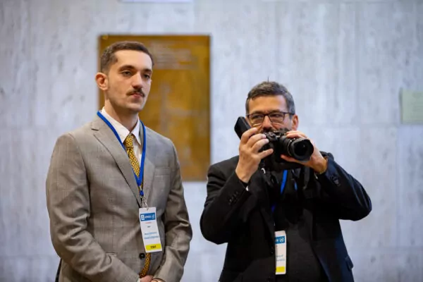 Two men wearing conference badges are standing indoors; one in a gray suit with a serious expression, the other in a black suit holding a camera and capturing this historic meeting for multilateralism. A marble wall and a plaque are visible in the background. - Rotary Club Paris Agora