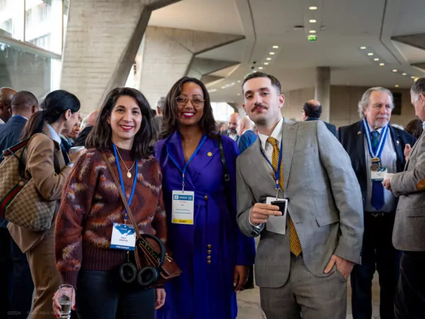 A group of people at a professional event, with three people in the foreground wearing conference badges and posing for the camera; other people mingle in the background during a historic meeting on the future of multilateralism in a modern indoor setting. - Rotary Club Paris Agora