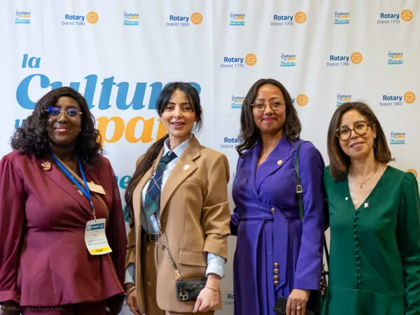 Four women stand side-by-side, smiling at the camera in front of a backdrop adorned with Rotary logos and the phrase "Culture," marking a historic moment for multilateralism. They wear work attire with badges and pins. - Rotary Club Paris Agora