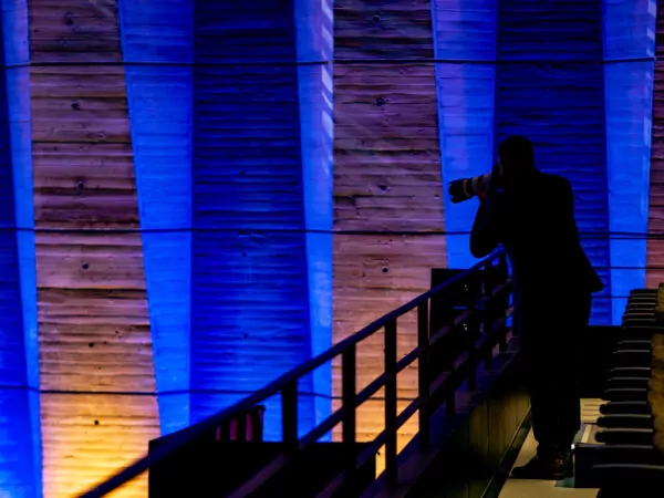 A person with a camera and a telephoto lens stands on a balcony, their silhouette set against blue and orange illuminated wall panels – an atmosphere evoking a historic meeting. Empty seats occupy the foreground, awaiting a future shaped by captured moments. - Rotary Club Paris Agora