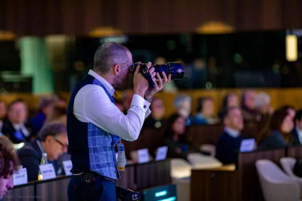 A man wearing a shirt and vest takes photos with a professional camera at an indoor event, capturing a historic meeting as seated participants watch and listen in the background. - Rotary Club Paris Agora