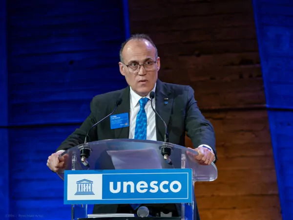 A man wearing glasses and a suit speaks on a brightly lit stage with a blue UNESCO sign, holding documents, against a wooden background, evoking the future of multilateralism on the occasion of this historic meeting. - Rotary Club Paris Agora