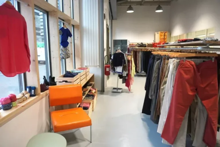Intérieur d'un magasin de vêtements avec des étagères de vêtements, une chaise orange, une petite table ronde, des étagères avec des articles pliés et du fil pour le textile de recyclage, de grandes fenêtres sur la gauche et des plafonniers. - Rotary Club Paris Agora