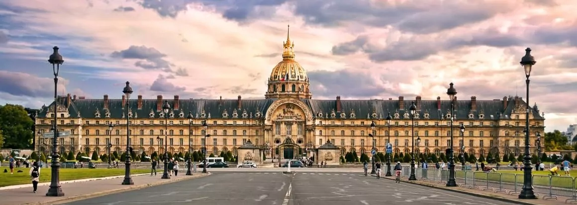 Vue grand angle de l'Institution nationale des Invalides à Paris, montrant la grande façade et le dôme central sous un ciel partiellement nuageux. Des lampadaires encadrent la scène tandis que des personnes marchent et se rassemblent au premier plan. - Rotary Club Paris Agora
