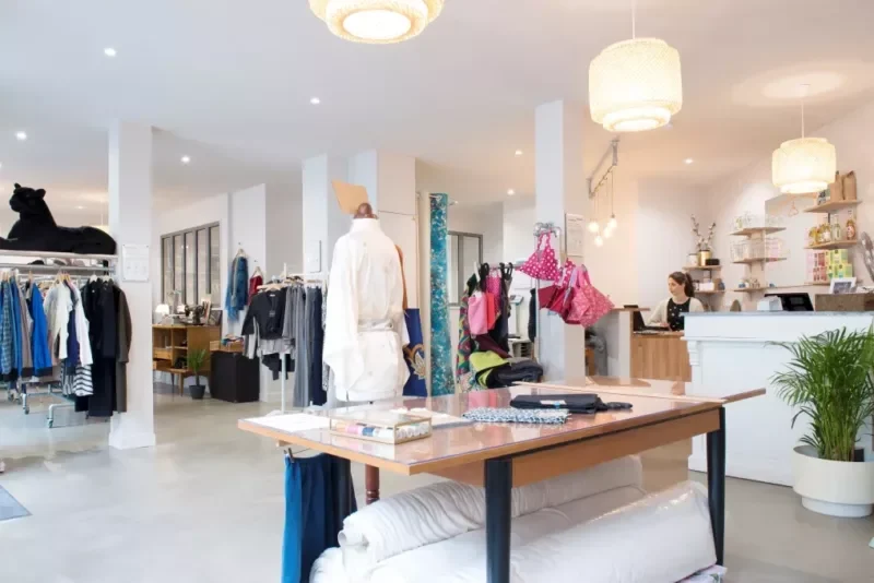 Une boutique de vêtements moderne présentant des étagères de vêtements, un mannequin, des sacs à main et des textiles pliés. Le décor minimaliste du magasin fait également la promotion du textile recyclé, encourageant les clients à participer à la mode durable. - Rotary Club Paris Agora