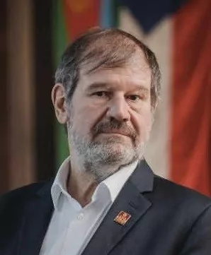 Un homme d'âge moyen aux cheveux et à la barbe grisonnants, vêtu d'une chemise blanche et d'une veste sombre, se tient devant des drapeaux aux couleurs floues lors d'une manifestation de sensibilisation à la polio. - Rotary Club Paris Agora