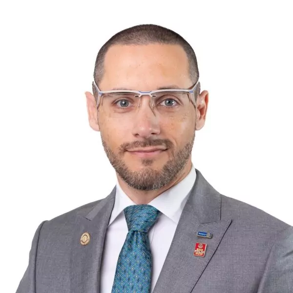 Un homme aux cheveux courts, barbu et portant des lunettes est vêtu d'un costume gris, d'une chemise blanche et d'une cravate à motifs sarcelle. Il porte deux épingles à la boutonnière et fait face à la caméra sur un fond blanc. - Rotary Club Paris Agora