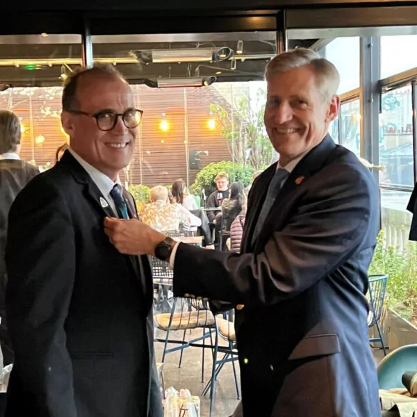 Deux hommes en costume se tiennent à l'intérieur ; l'un épingle un badge sur le revers de l'autre en souriant, célébrant un moment important dans l'histoire du club. Des personnes sont assises à des tables et des lampes suspendues sont visibles à l'arrière-plan. - Rotary Club Paris Agora