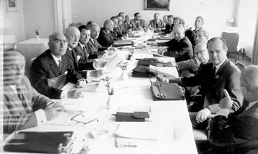 Black and white photo showing a group of men in suits sitting around a long table, participating in an Intercountry Committee meeting. Papers, folders, and documents are spread across the table in a formal office setting. - Rotary Club Paris Agora