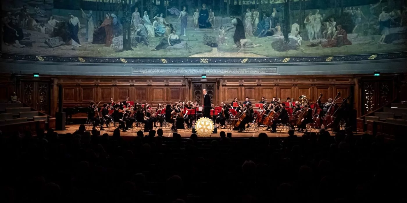 Un orchestre donne un concert sur la scène d'une grande salle lambrissée et surmontée d'une peinture murale. Les musiciens jouent de divers instruments tandis qu'un chef d'orchestre se tient au centre, face à l'orchestre, évoquant la grandeur de la Sorbonne. - Rotary Club Paris Agora