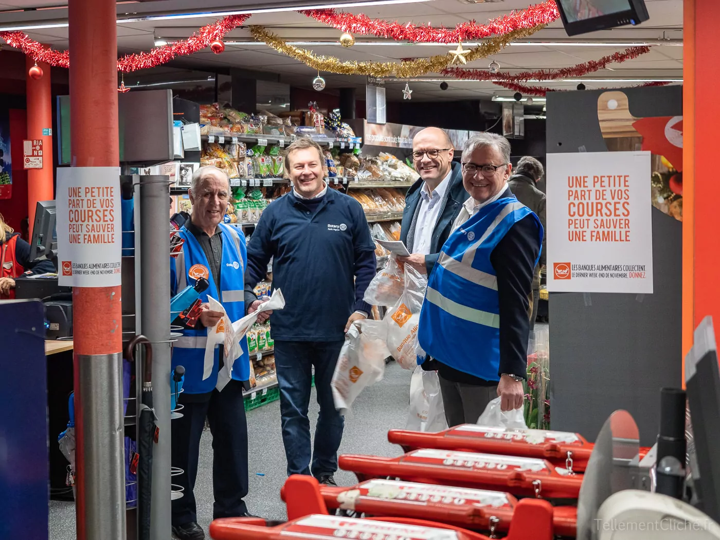 Quatre hommes sourient à l'intérieur d'un magasin d'alimentation près de la caisse, deux d'entre eux portent des gilets bleus. On aperçoit à l'arrière-plan des étagères de fruits et légumes, des décorations de Noël et des panneaux de la Banque Alimentaire collecte en français. - Rotary Club Paris Agora